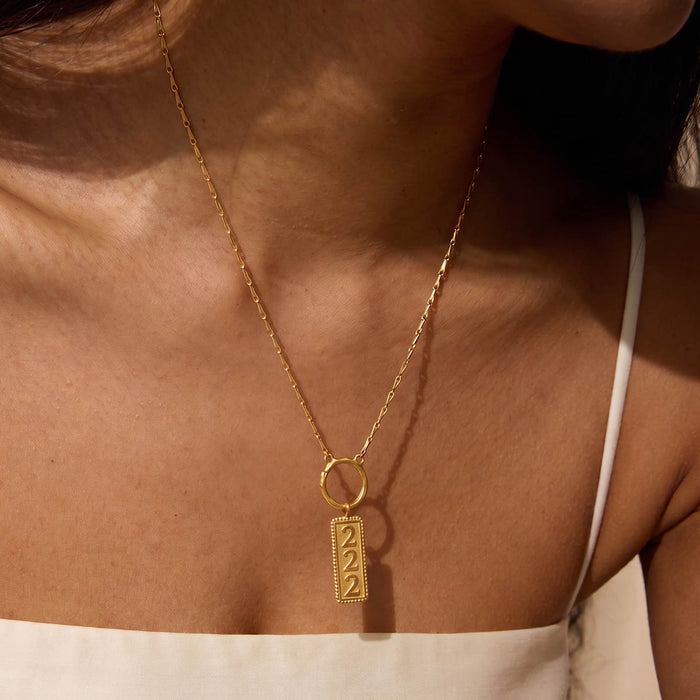 Gold-toned necklace with rectangular charm engraved with "222" worn close to chest.