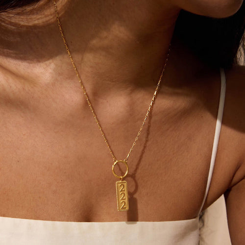 Gold-toned necklace with rectangular charm engraved with "222" worn close to chest.