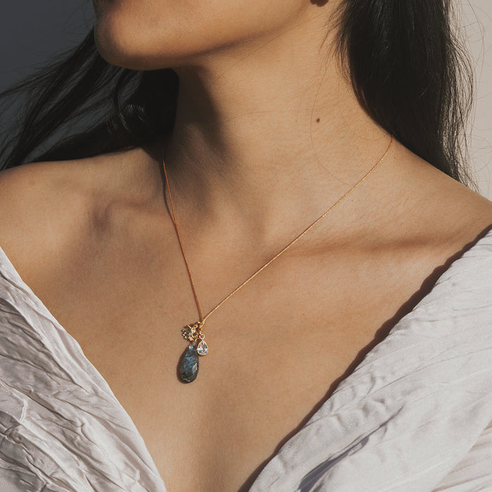 Thin cord necklace with gray labradorite stone pendant and gold-toned lotus charm accent worn close to collarbone.