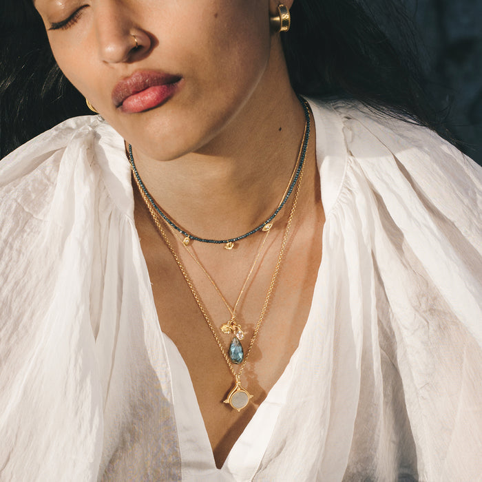 Layered gold chain necklaces with iridescent labradorite lotus charm and clear stone pendant worn on model in white shirt.