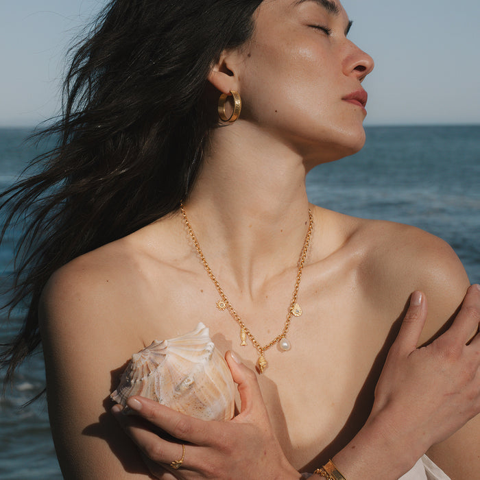 Gold chain necklace with pearl and shell-shaped charm pendant worn by woman at ocean.