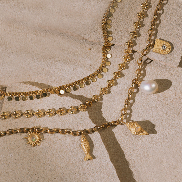 Gold-toned layered chains with pearl, fish, sun, and shell charms on sand background.
