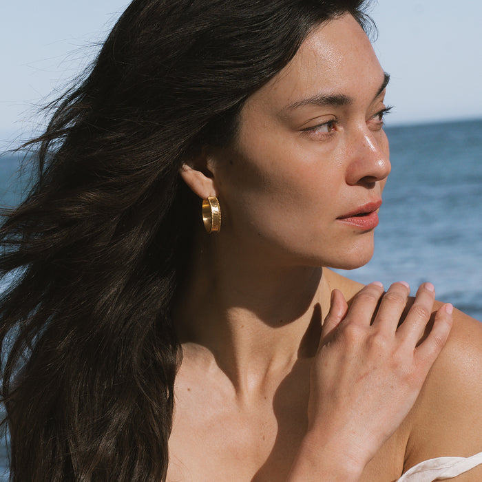 Gold hoop earrings with textured surface worn by person with long dark hair.