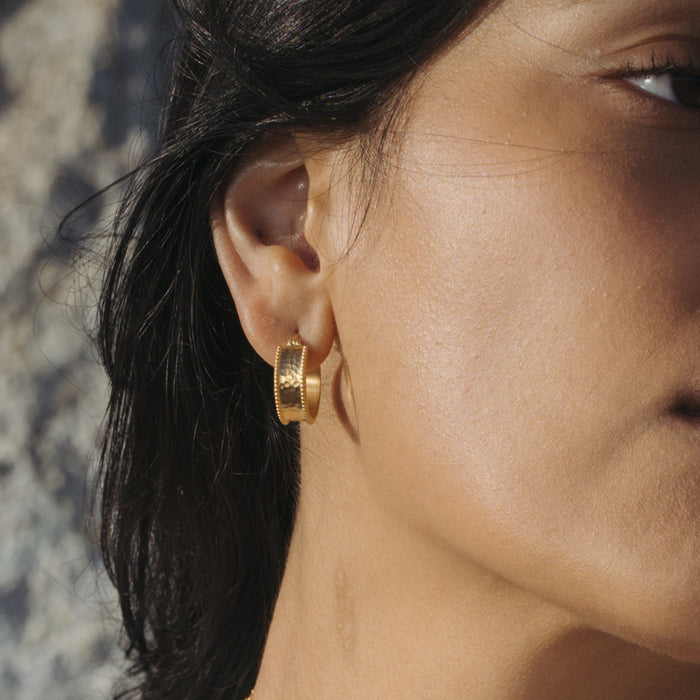Gold-toned rectangular hoop earrings with textured surface worn on a person's ears.