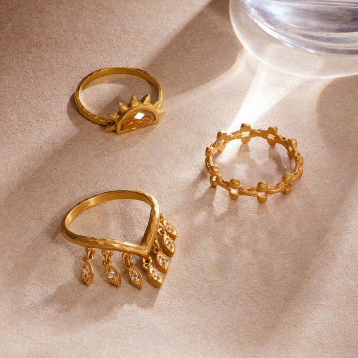 Three gold-toned rings with petal and geometric designs displayed on a beige surface.