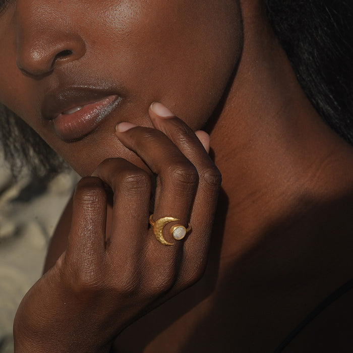 Gold-toned ring with pale moonstone stone worn on ring finger of person with deep skin tone.