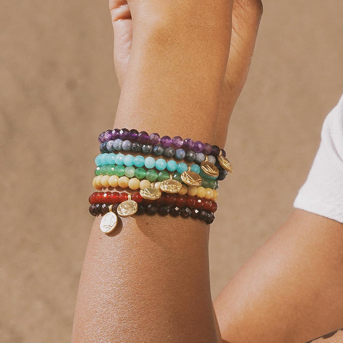 A collection of colorful stretch bracelets worn on a wrist, including the Crown Chakra Amethyst Stretch Bracelet. This assortment of spiritual jewelry symbolizes meditation, protection, and meaning, making it a perfect accessory for spiritual seekers and yoga practitioners. The combination of vibrant beads serves as a beautiful reminder of manifestation and the energy of the sun and moon.