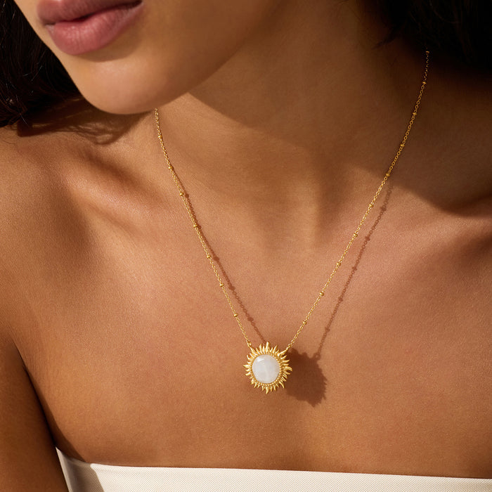 Gold-toned necklace with a sun-shaped pendant featuring a white moonstone center, worn on a person's chest.