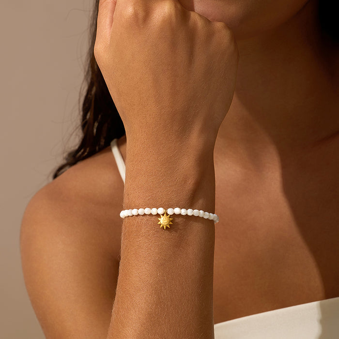 White mother of pearl beaded stretch bracelet with small gold sun charm worn on wrist.