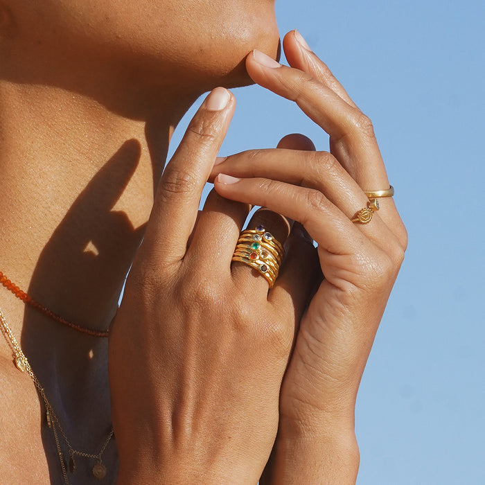 Gold stacked ring with colorful gemstone accents worn on model's hand against blue sky.