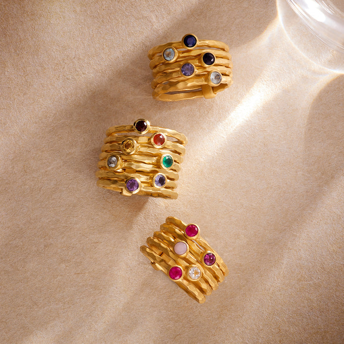 Three gold stacked rings with colorful gemstones in purple, blue, red, green, and clear settings.