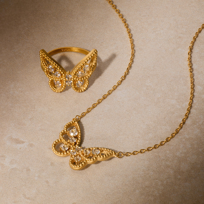 Gold butterfly necklace and ring set with white topaz stones on textured background.