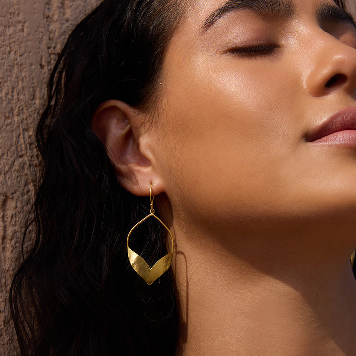 Gold teardrop earrings with pointed petal-shaped center worn on model against textured brown wall.