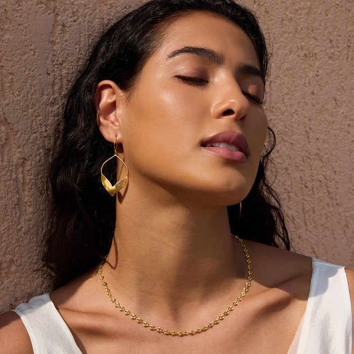 Gold petal-shaped drop earrings with curved design worn on person against textured beige background.