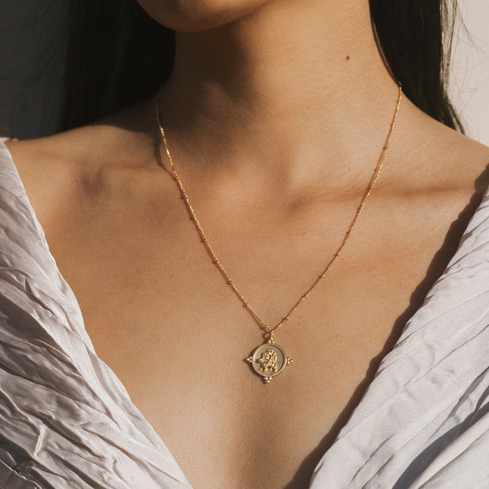 Gold-toned necklace with circular coin pendant featuring ornate deity design, worn on woman's chest.