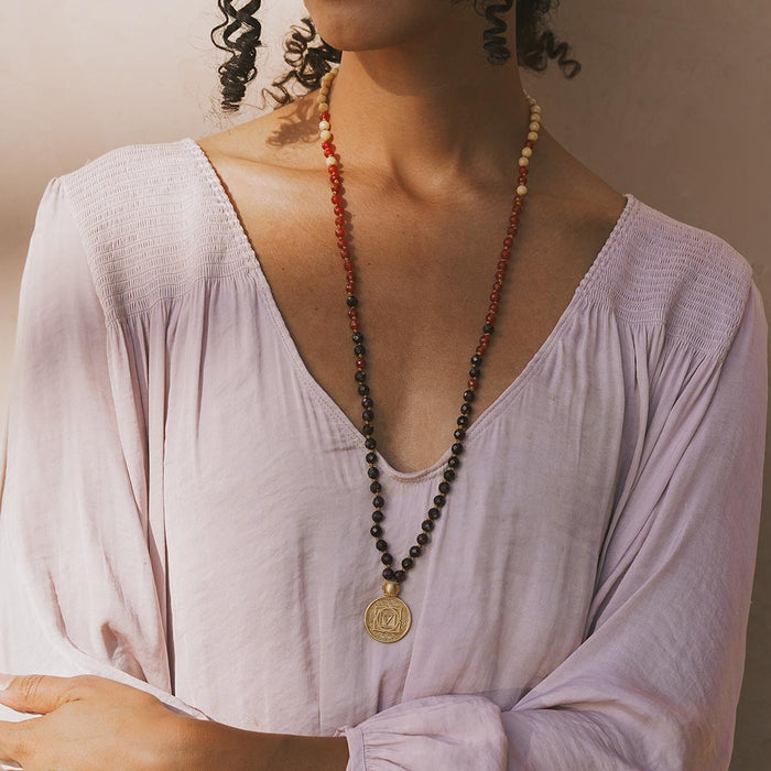 Long beaded mala necklace with red, black, and natural stone beads, gold circular pendant, worn over lavender top.
