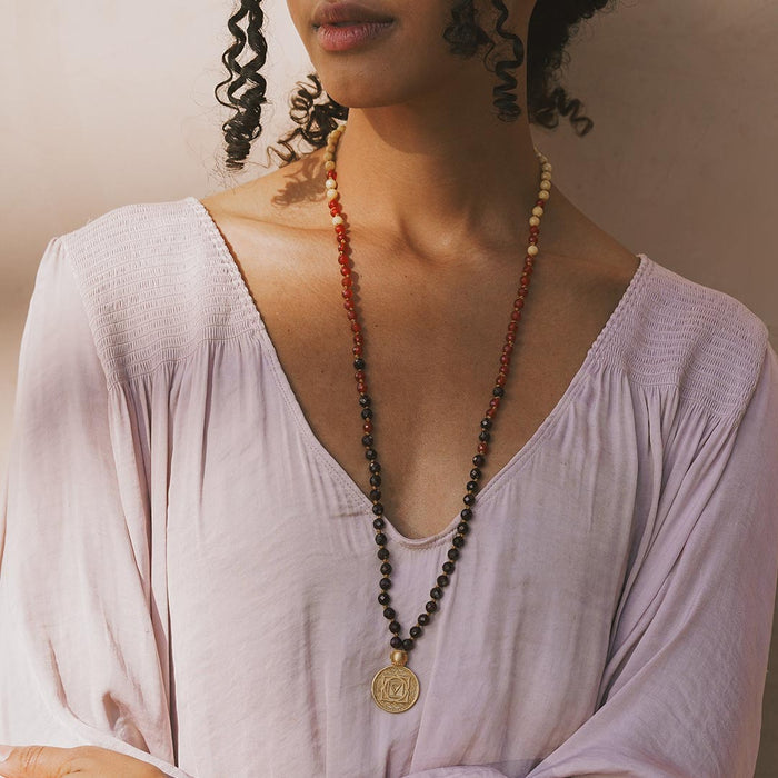 Beaded mala necklace with red and black stones, cream beads, and gold pendant charm worn long across chest.