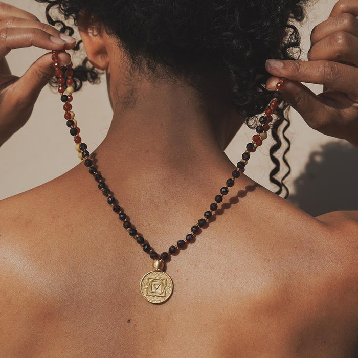 Beaded mala necklace with dark red and black stones and gold medallion pendant worn around person's neck.