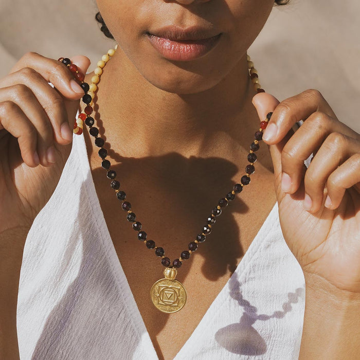 Beaded mala necklace with black and gold faceted beads, gold chakra pendant, and cream-colored tassel worn on person.