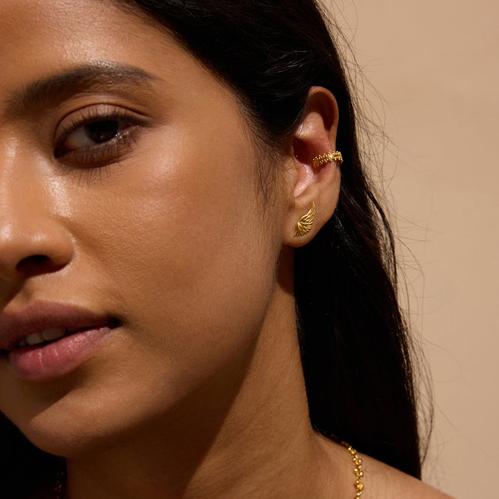 Gold-toned wing-shaped stud earrings worn on woman's earlobe against neutral background.