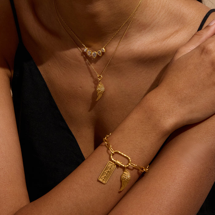 Gold necklace with angel wing pendant and matching bracelet with wing charm worn on model's neck and wrist.