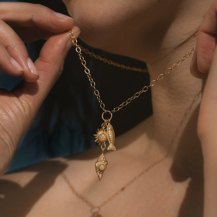 Gold-toned necklace with shell and fish charms featuring pearl accents, worn on a person's chest.