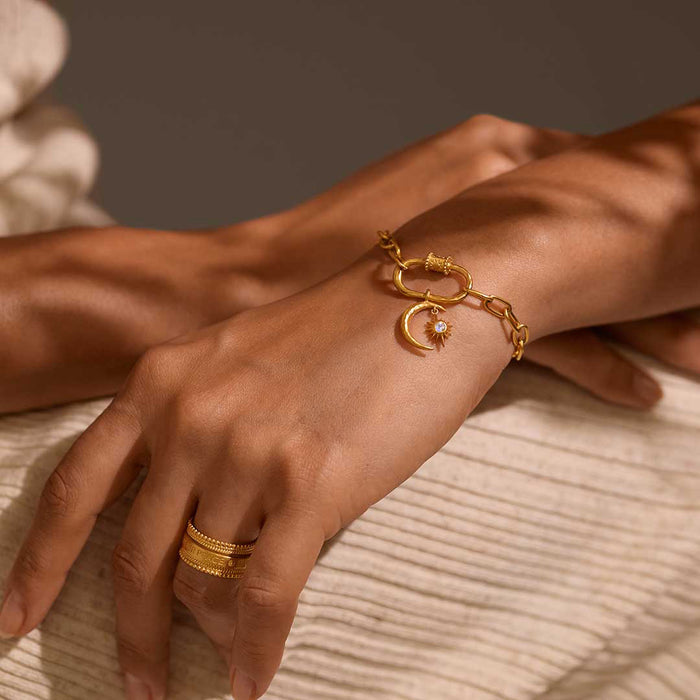 A close-up of a hand wearing the Infinite Possibilities Moonstone Celestial Charm on a bracelet, showcasing its 18kt gold plating and crescent moon design. This spiritual jewelry piece symbolizes meditation and manifestation, making it a meaningful talisman for those who seek protection and guidance from the sun and moon in their yoga practice.