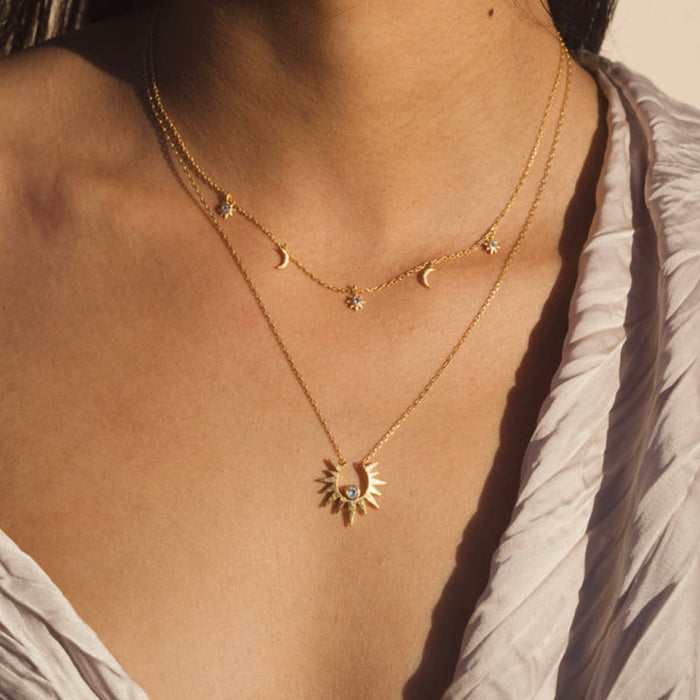 Two layered gold necklaces with moon, star, and sun pendants worn on a person's chest.