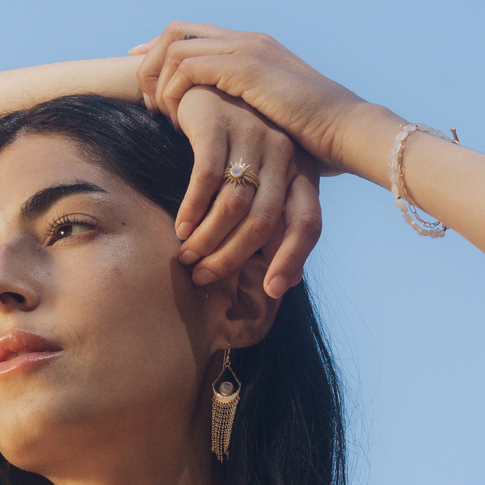 A woman showcases the Light Seeker Moonstone Starburst Nested Ring Set while holding her hair, representing spiritual jewelry that embodies clarity and protection. This piece, adorned with celestial charm, signifies meditation, manifestation, and the connection to the sun and moon, making it a meaningful talisman for yoga and zodiac enthusiasts.