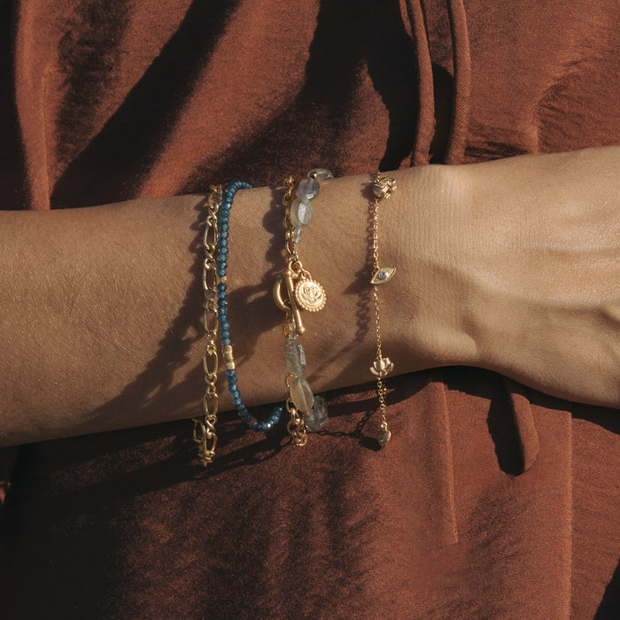 Gold-toned chain bracelet with blue beads, gray stone accents, and circular pendant charm worn on wrist.