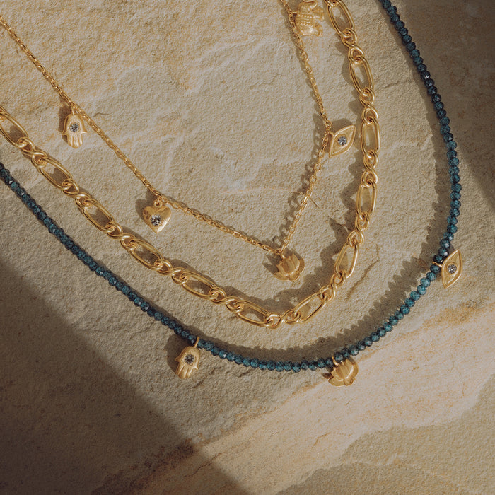 Three layered chain necklaces in gold and blue with small pendant charms on textured background.