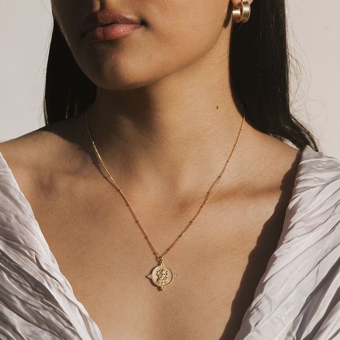 Gold-toned necklace with circular coin pendant featuring deity figure, worn on model's chest.
