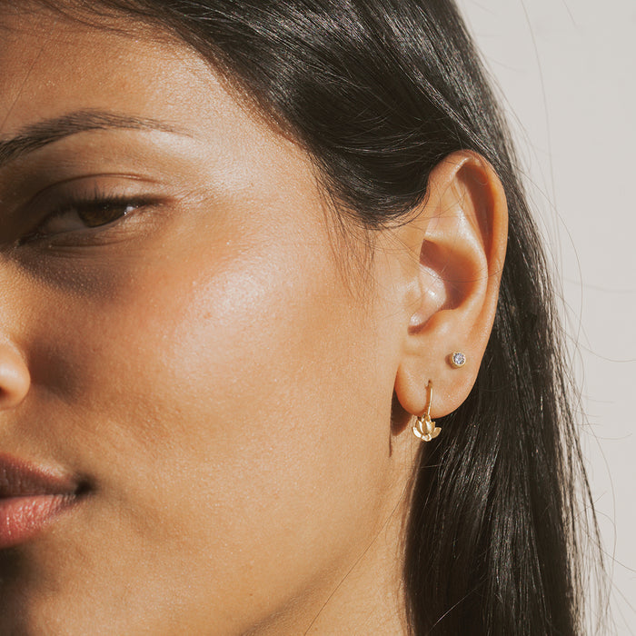 Pair of gold-toned lotus-shaped stud and hoop earrings with white topaz stones worn on earlobe.