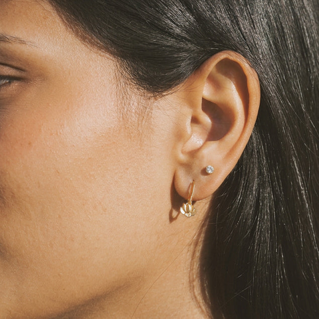 Person wearing a pair of small round clear gemstone stud earrings and small metallic hoop earrings on lower lobe piercings.