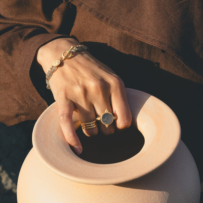 Gold-tone adjustable ring with clear round stone worn on hand with bracelet and brown fabric.