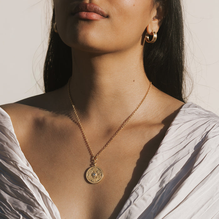 Gold-toned circular coin pendant with geometric design hanging from delicate chain necklace worn on model.
