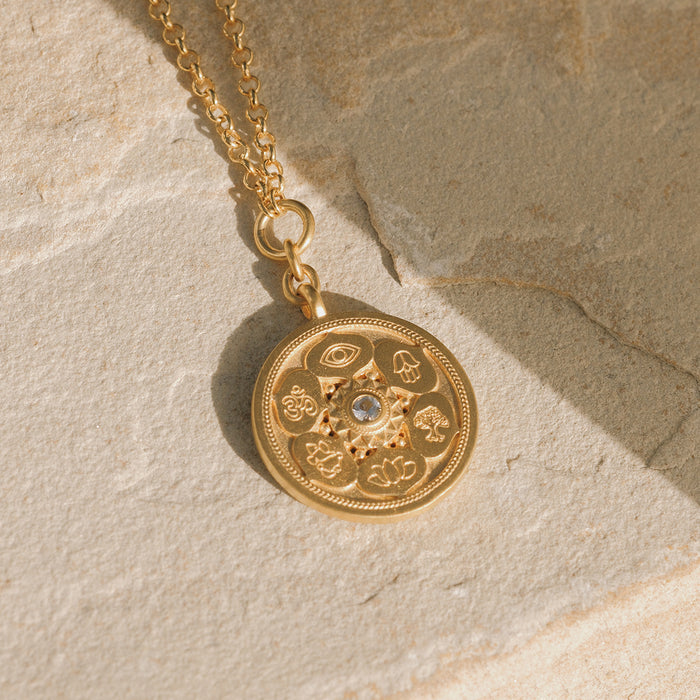 Gold coin pendant necklace with etched spiritual symbols including an eye, Om, hamsa, and other religious icons arranged in a