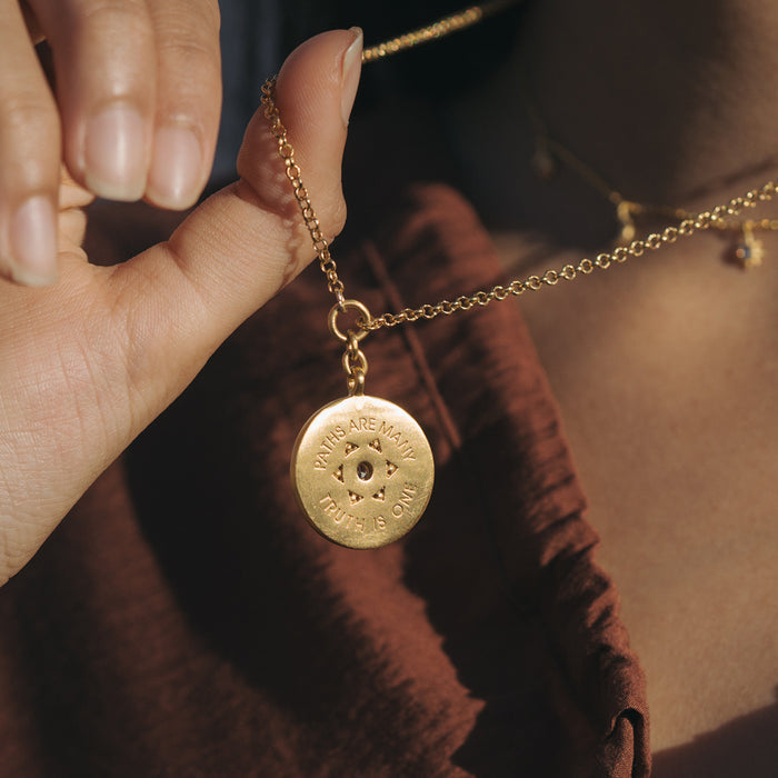 Gold-toned coin pendant necklace with engraved text and sun symbol, held in hand on gold chain.