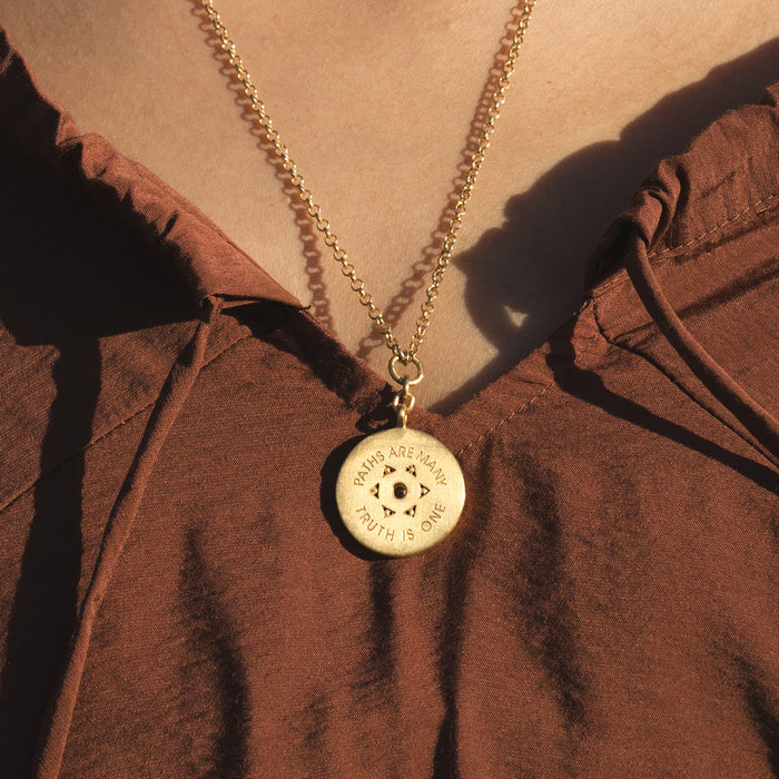 Gold-tone necklace with circular pendant featuring engraved text and eye symbol, worn on brown fabric.