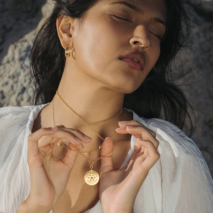 Gold chain necklace with circular coin pendant featuring geometric pattern, worn by person in white shirt.
