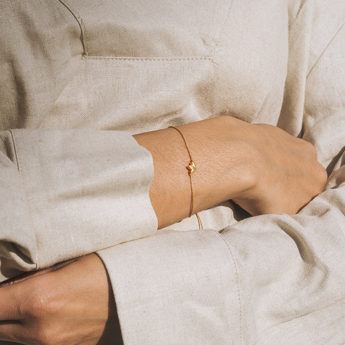 Tan cord bracelet with small gold lotus charm worn on wrist against beige fabric.