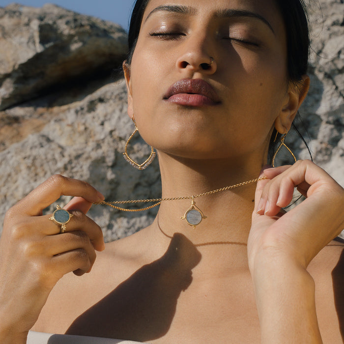 Gold-toned chain necklace with round labradorite pendant and matching ring worn by model outdoors.