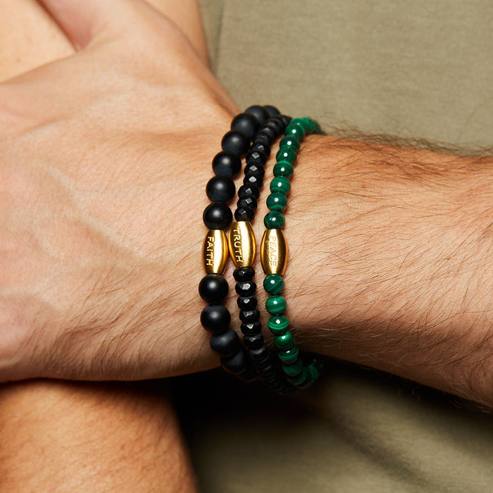 A stylish wrist adorned with the Path to Healing Malachite Gemstone Men's Bracelet alongside black and green beaded bracelets, showcasing a blend of spiritual jewelry and talismans for yoga practices. Perfect for those seeking harmony, meaning, and charm in their accessories.