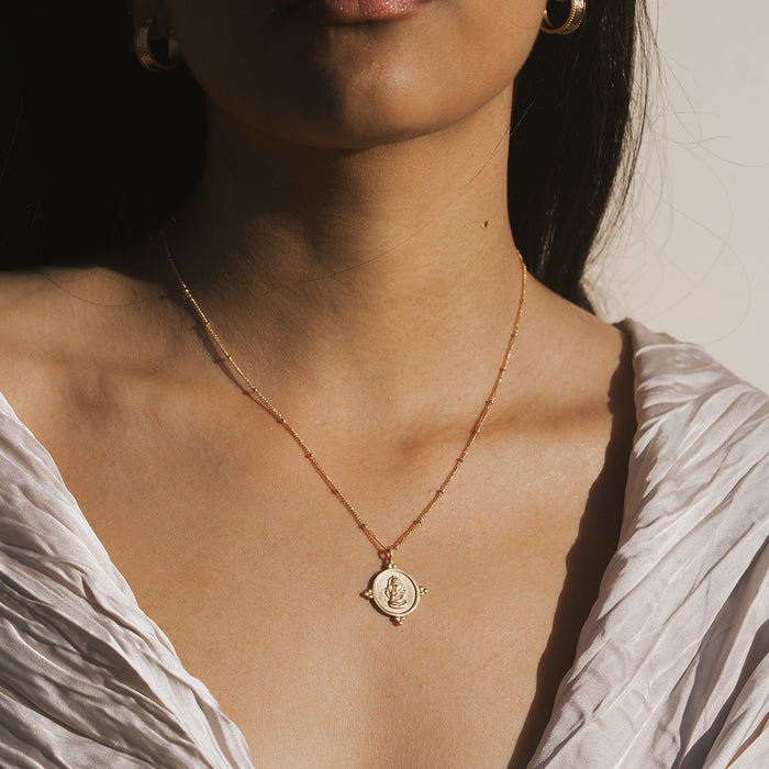 Gold-toned necklace with round pendant featuring Ganesha deity design, worn on model's chest.