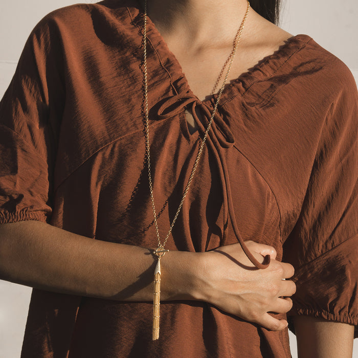 Gold-tone chain necklace with tassel pendant worn long over brown textured blouse.