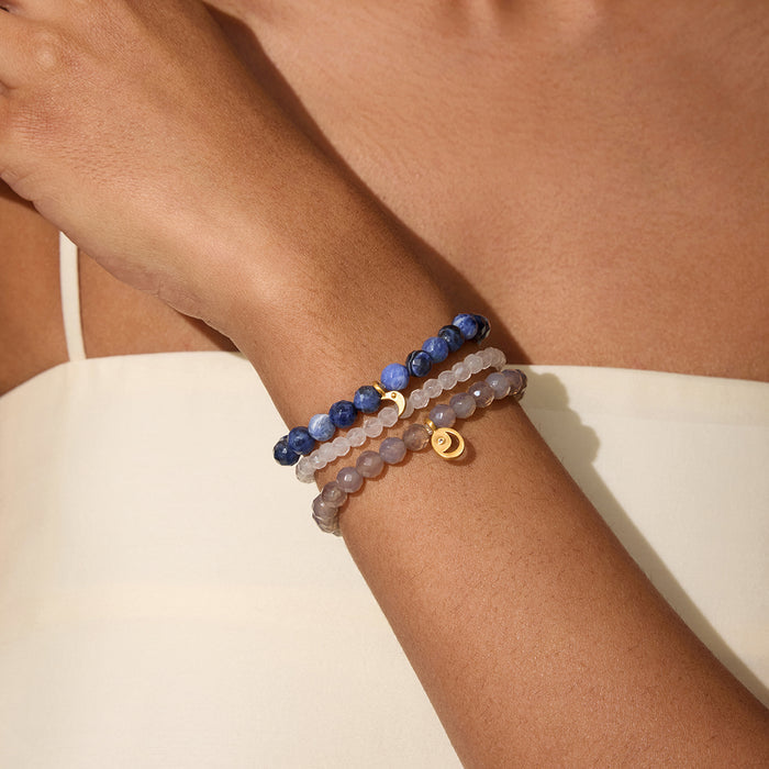 Three beaded bracelets stacked on wrist: dark blue sodalite, light rose quartz, and gray gemstone strands with gold moon char
