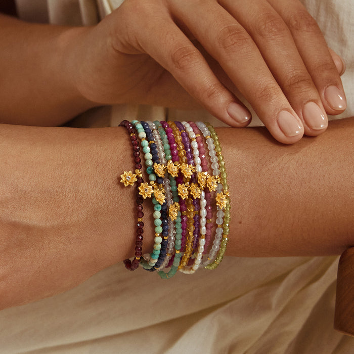 Beaded bracelet stack with red, purple, white, and green stones and gold lotus flower charms worn on wrist.