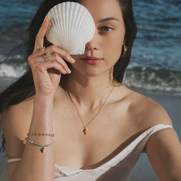Gold-toned necklace with small opal shell pendant worn by woman at beach holding white conch shell.
