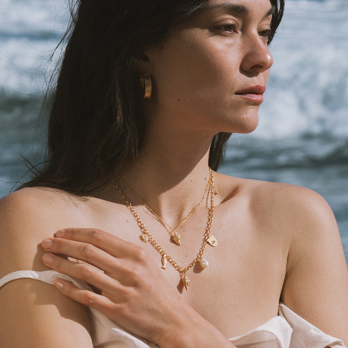 Gold-toned layered necklace with iridescent opal conch shell pendants worn by a woman against an ocean backdrop.