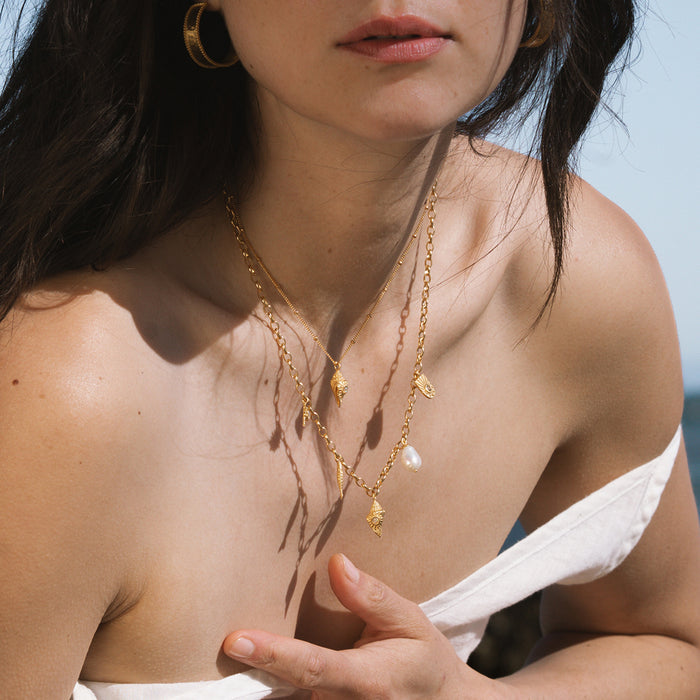 Gold chain necklace with small white opal and shell charms, worn layered on a person's chest.